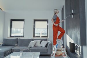 A woman on a ladder cleaning the wall of a grey-white living room.